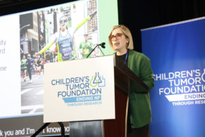 Woman in glasses and green cardigan speaking at a podium with the children’s tumor foundation logo, with a marathon image in the background.