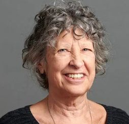 Portrait of an elderly woman with curly gray hair smiling, wearing a dark sweater against a gray background.