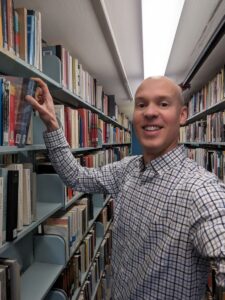 A man in a plaid shirt smiling and selecting a book from a shelf in a library aisle.