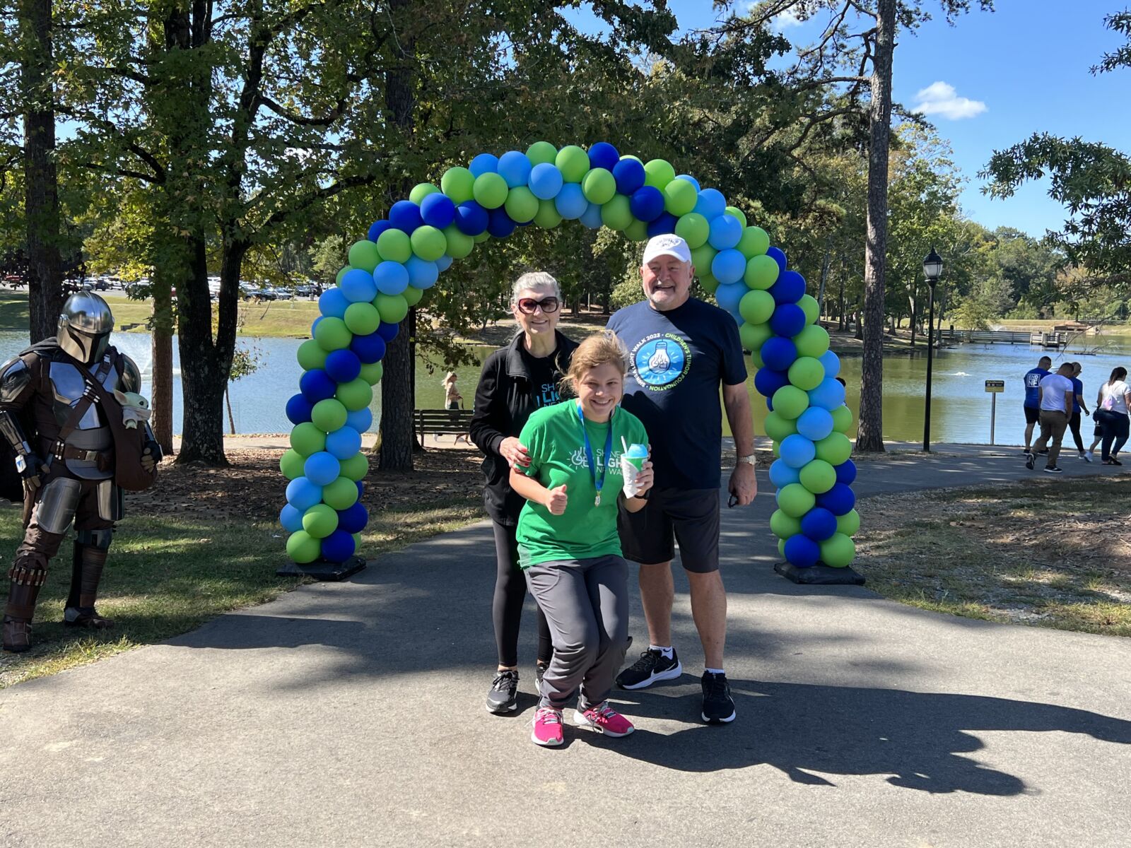 Family poses with a costumed character under a blue and green balloon arch in a park.