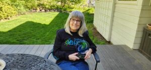 Elderly woman with glasses and long gray hair sitting outdoors, holding a blue glass award, wearing a black hoodie with colorful text.