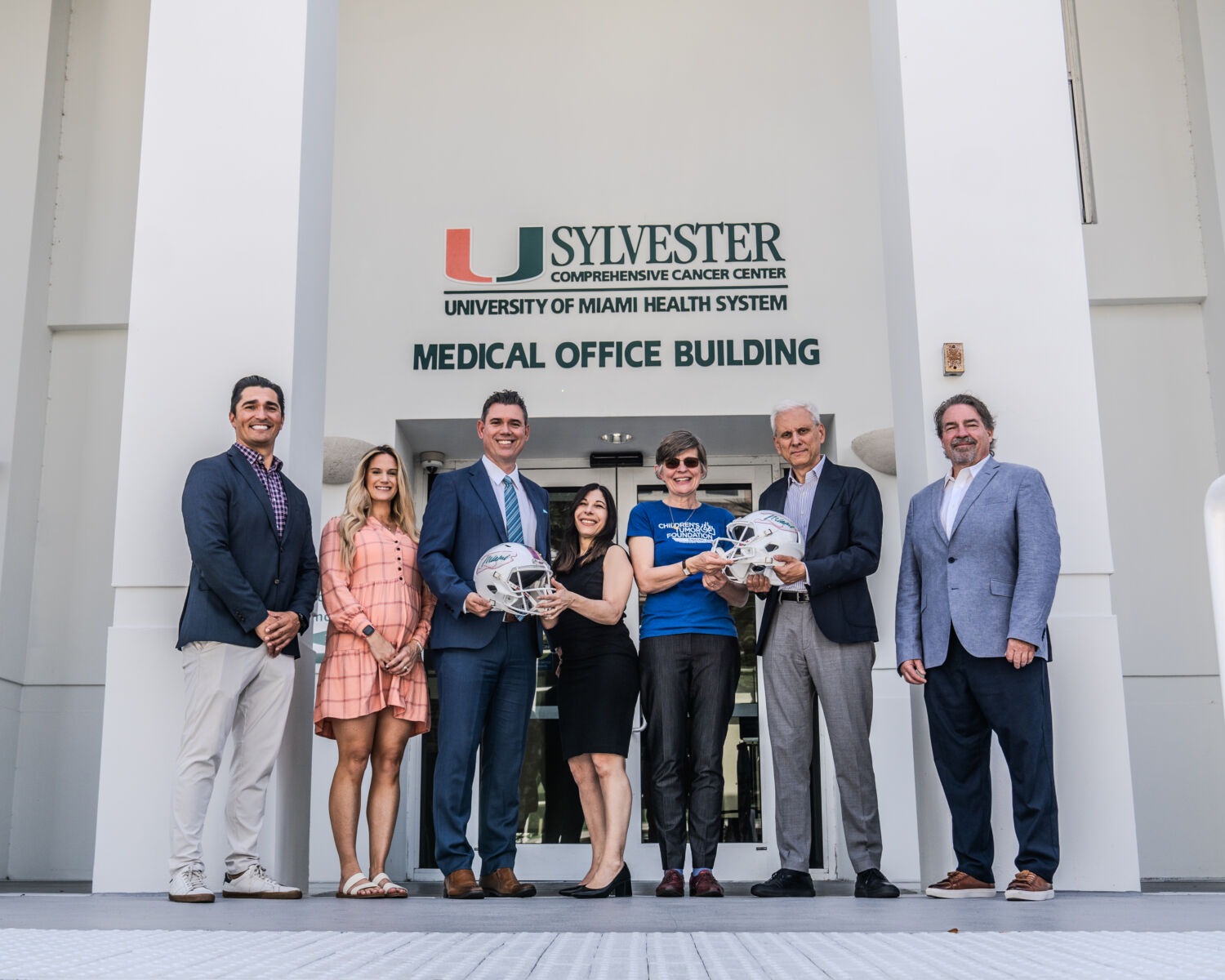 Group of seven adults standing in front of the university of miami sylvester comprehensive cancer center, holding commemorative football helmets.