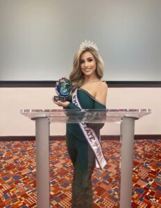 A woman in a green dress and a sash stands behind a podium, holding a trophy and wearing a tiara, smiling in a conference room.