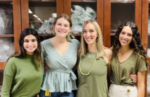 Four smiling people stand arm-in-arm in front of wooden cabinets filled with plastic bags. Three of them are wearing green tops, while one is wearing a light blue floral top.