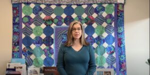 A woman with glasses wearing a blue sweater stands in front of a colorful quilt hanging on a wall.