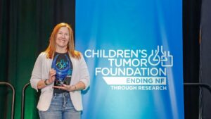 Woman smiling while holding an award at the children's tumor foundation event, standing in front of a banner displaying the foundation's logo.