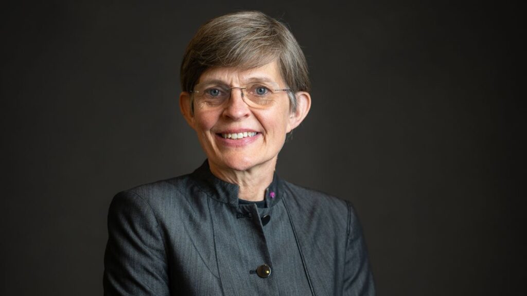 A person with short hair, glasses, and a black jacket smiles against a dark background.