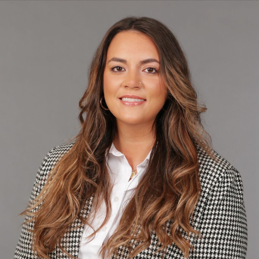 A person with long wavy hair, wearing a black and white checkered blazer and a white shirt, against a gray background.