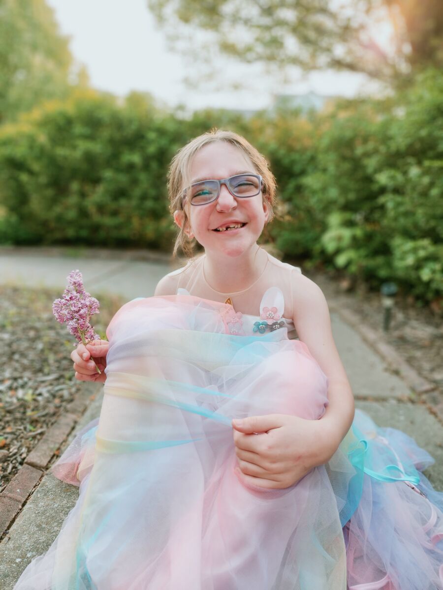 A child in glasses smiles while holding a purple flower, wearing a colorful dress. They are outdoors with greenery in the background.