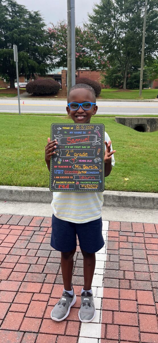Child holding a colorful first day of school sign while standing outdoors on a brick path, with grass and trees in the background.