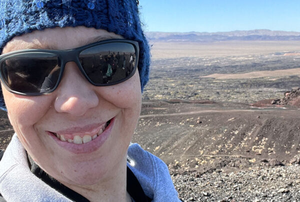 Person wearing sunglasses and a blue beanie smiling at the camera with a rocky landscape and distant mountains in the background.