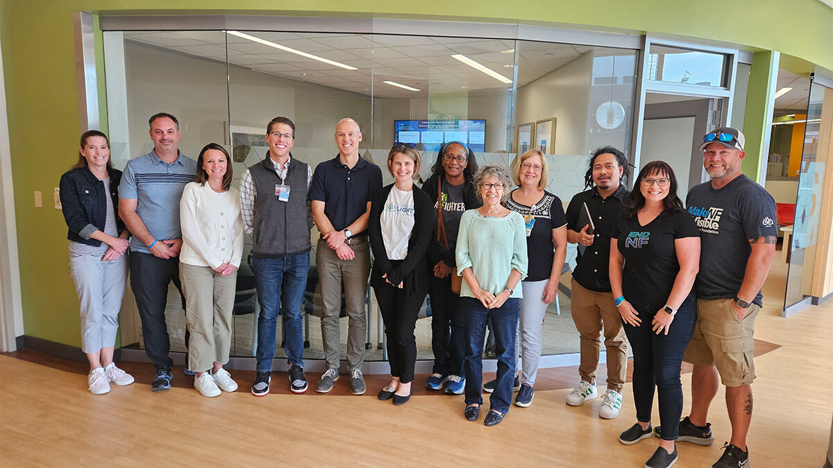 A group of 12 people stands in a row inside a building with glass walls. Some are wearing casual attire, and all are smiling.