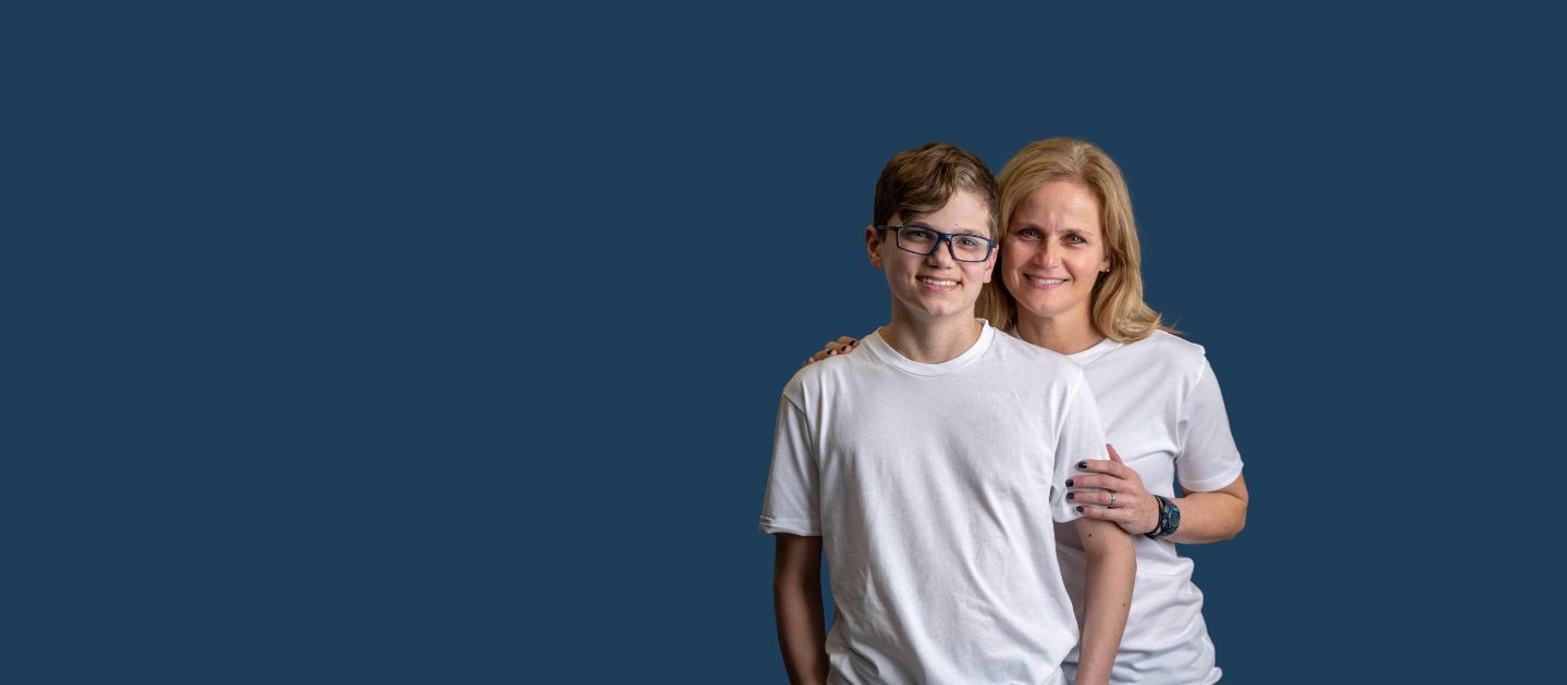 A woman and a boy wearing white t-shirts stand arm-in-arm against a solid blue background.