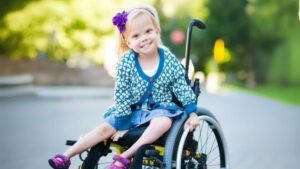 A young girl with a flower in her hair sits smiling in a wheelchair on a sunny day, wearing a patterned cardigan and a denim skirt.