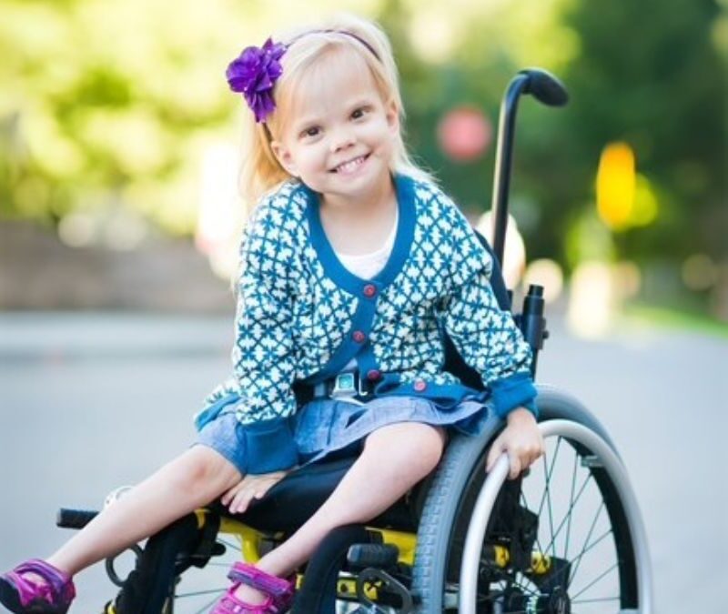 A young girl with a flower in her hair sits smiling in a wheelchair on a sunny day, wearing a patterned cardigan and a denim skirt.