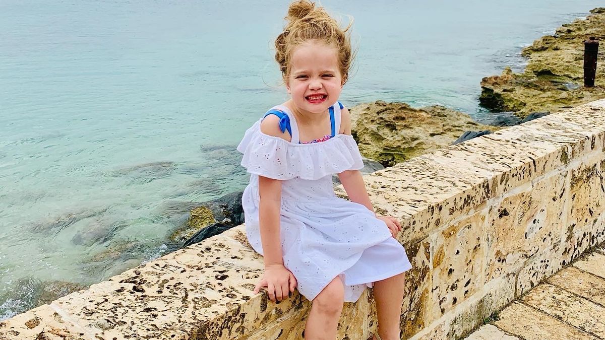 Child in a white dress sits on a stone wall by the sea, smiling, with a rocky shoreline and turquoise water in the background.