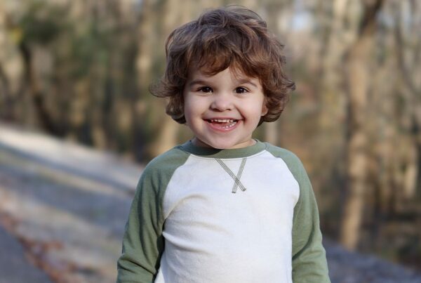 A young child with curly brown hair and a green and white shirt smiles outdoors with a blurred forest background.