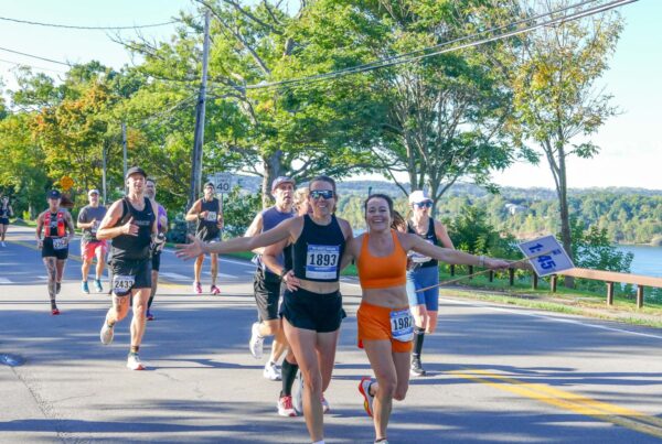 Two smiling runners in athletic gear embrace while running on a sunny road with other participants. Trees and water are visible in the background.