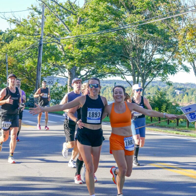 Two smiling runners in athletic gear embrace while running on a sunny road with other participants. Trees and water are visible in the background.