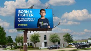 A roadside billboard displays a young boy with the text, “Born a Fighter. He shouldn’t have to fight tumors alone.” and website endnf.org against a blue background.