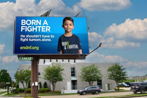 A roadside billboard displays a young boy with the text, “Born a Fighter. He shouldn’t have to fight tumors alone.” and website endnf.org against a blue background.
