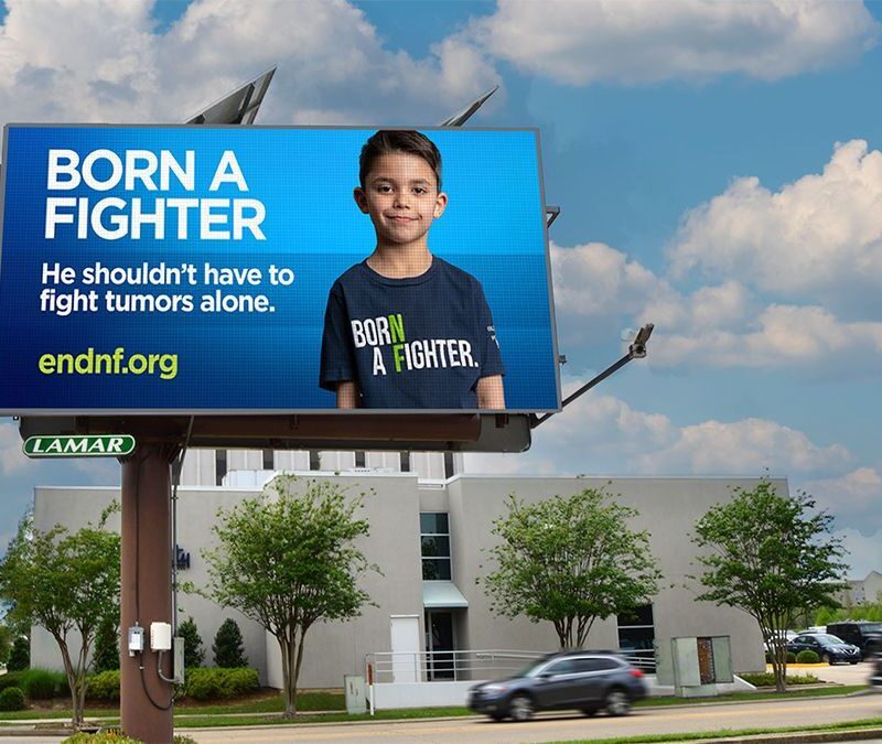 A roadside billboard displays a young boy with the text, “Born a Fighter. He shouldn’t have to fight tumors alone.” and website endnf.org against a blue background.