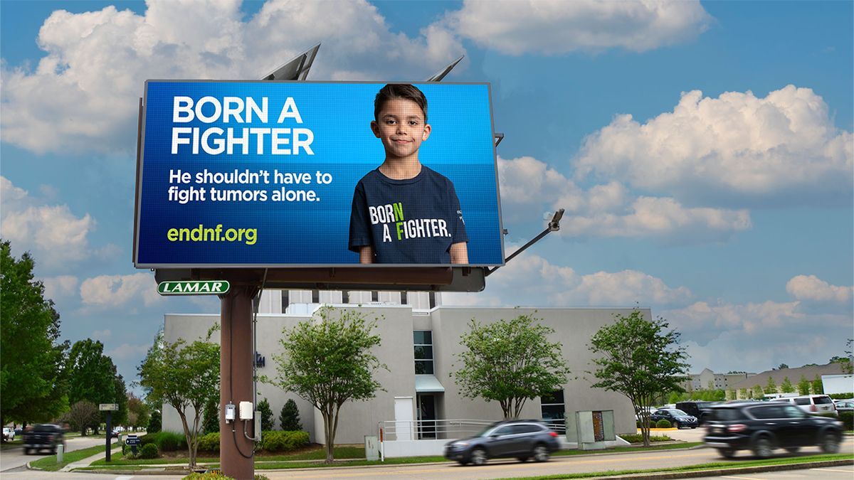 A roadside billboard displays a young boy with the text, “Born a Fighter. He shouldn’t have to fight tumors alone.” and website endnf.org against a blue background.