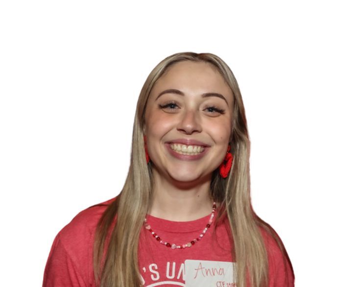 Anna Endsley A woman with long blonde hair smiles at the camera, wearing a red shirt, red earrings, a beaded necklace, and a name tag that reads "Anna." The background is white.