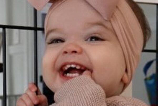 A smiling baby wearing a pink headband with a large bow and a knitted pink sweater, standing in front of a black metal gate.