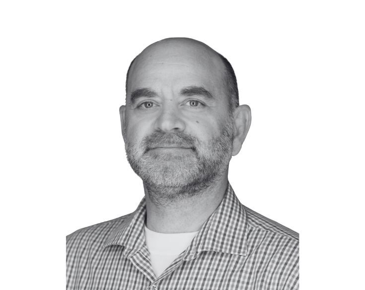 A middle-aged man with a beard and short hair wearing a checked shirt looks at the camera against a plain white background.