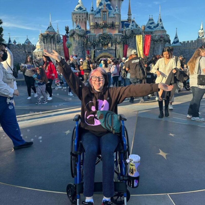 Person in a wheelchair smiling with arms raised in front of a castle at a theme park, surrounded by other visitors on a sunny day.