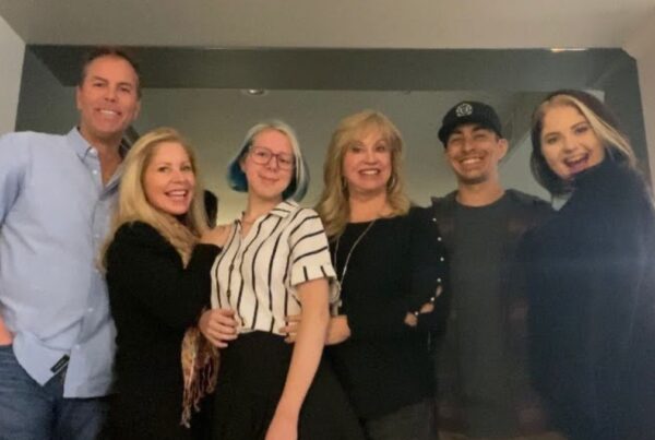 Six people stand closely together indoors, smiling at the camera in a kitchen or dining area.