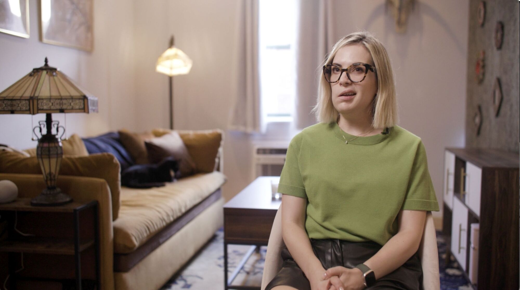 A woman wearing glasses and a green shirt sits on a chair in a living room with a brown sofa and a lamp in the background.