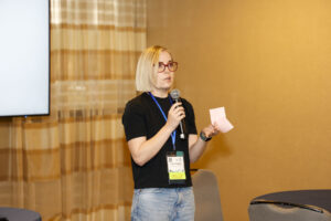 A person wearing glasses and a conference badge holds a microphone and a pink note while speaking in a conference room.