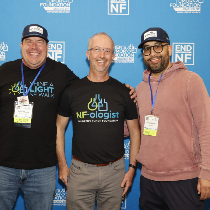 Three men stand smiling in front of a blue Children's Tumor Foundation backdrop, wearing event badges and foundation-themed shirts and hats.