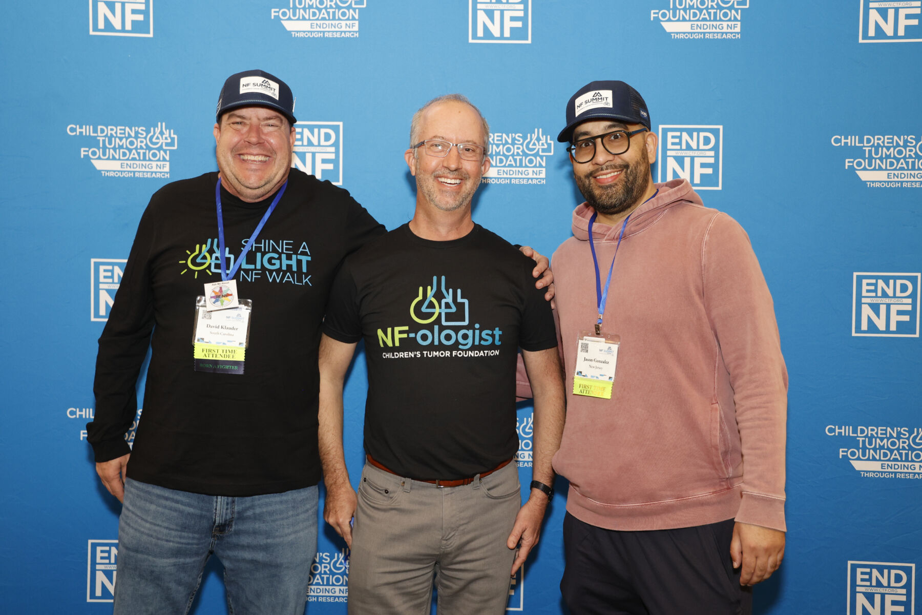 Three men stand smiling in front of a blue Children's Tumor Foundation backdrop, wearing event badges and foundation-themed shirts and hats.