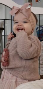 Smiling toddler wearing a pink headband and knitted sweater stands near a white crib, holding one hand to their mouth.