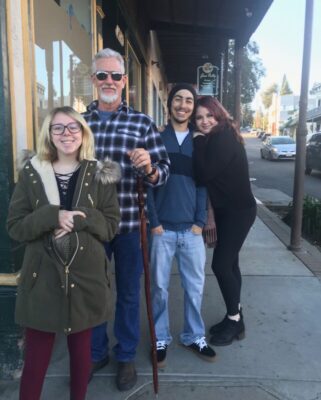 Four people stand together on a sidewalk in front of a building on a sunny day, smiling at the camera.