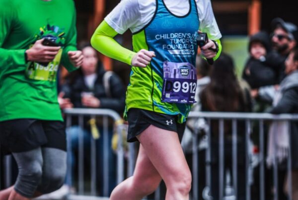 Runner participates in a road race; the woman in front wears sunglasses, a blue and green shirt, and a race bib, while spectators watch behind metal barriers.