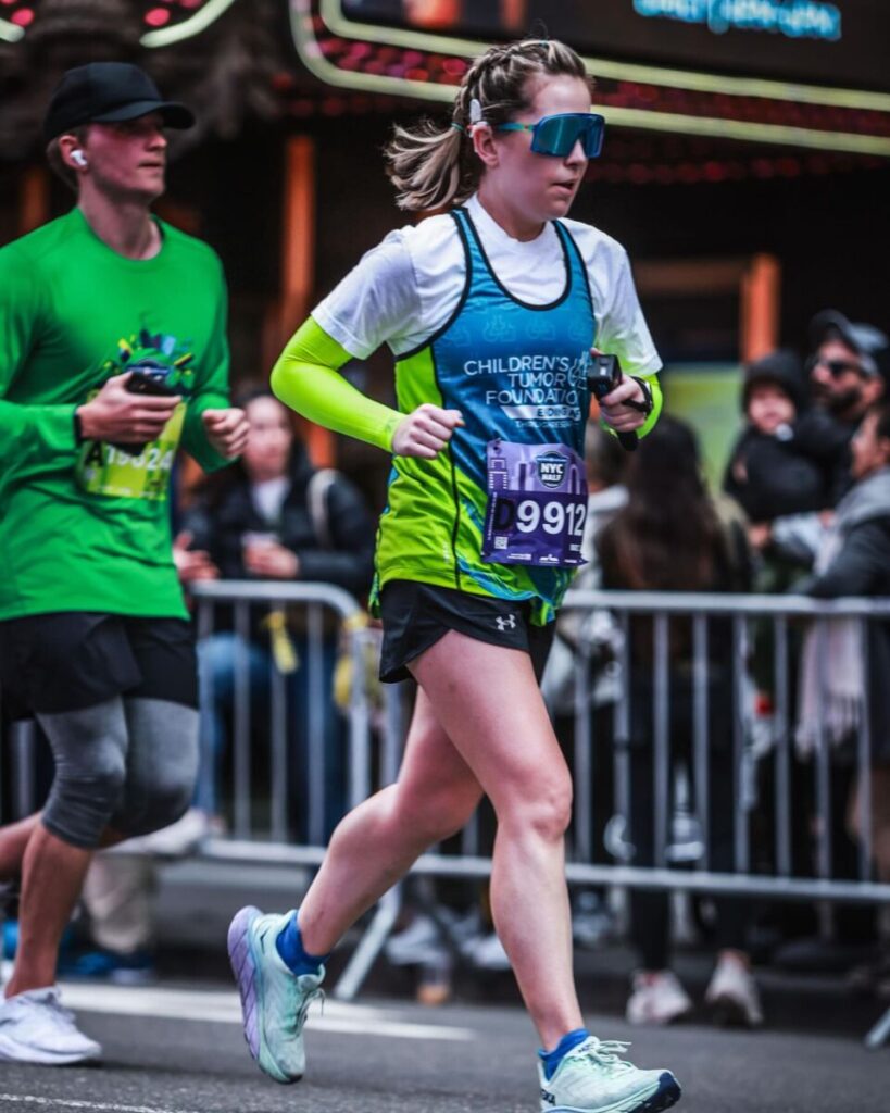 Runner participates in a road race; the woman in front wears sunglasses, a blue and green shirt, and a race bib, while spectators watch behind metal barriers.