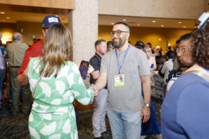 A man wearing glasses and a gray shirt shakes hands with a woman in a green and white dress at a busy indoor event.