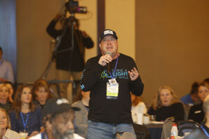 A man holding a microphone speaks during a conference while other attendees listen in the background.