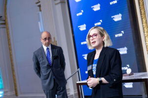 A woman speaks while a man stands in the background in a formal setting with an NYSE-branded screen behind them.