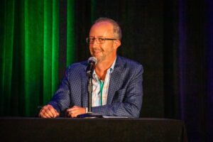 A man wearing glasses and a checked blazer sits at a table and speaks into a microphone against a dark background with green and black curtains.