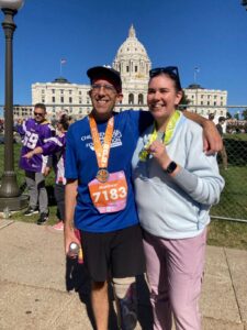 Two people stand smiling with medals after a race; the man wears a race bib and prosthetic leg, the woman wears sunglasses. A large capitol building is visible in the background.