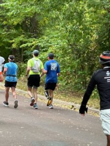 Four runners jog on a paved path through a wooded area. One runner wears a blue shirt with "END NF" on the back. The scene suggests an organized race or event.