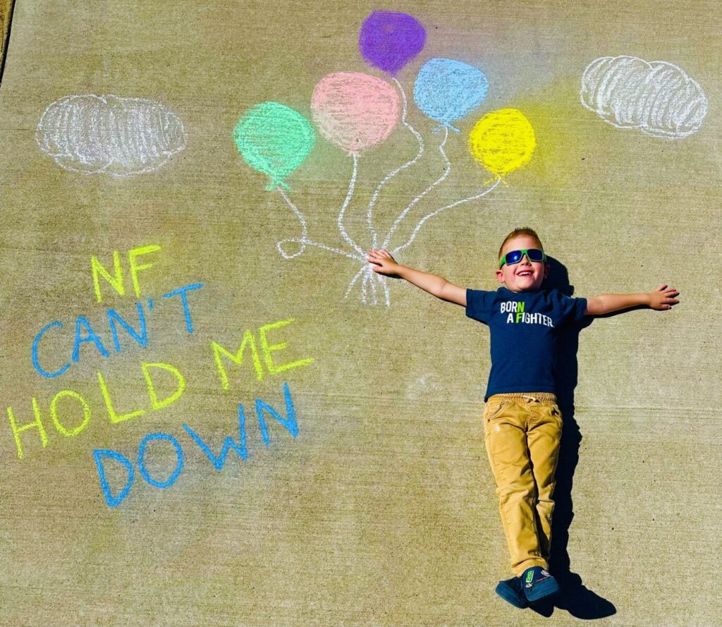 Young boy lies on pavement next to chalk drawings of balloons and clouds, with text reading "NF CAN'T HOLD ME DOWN." He wears sunglasses and a shirt that says "Born a fighter.