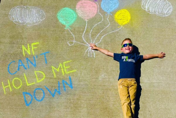 Young boy lies on pavement next to chalk drawings of balloons and clouds, with text reading "NF CAN'T HOLD ME DOWN." He wears sunglasses and a shirt that says "Born a fighter.