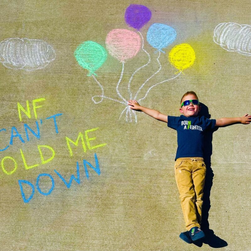 Young boy lies on pavement next to chalk drawings of balloons and clouds, with text reading "NF CAN'T HOLD ME DOWN." He wears sunglasses and a shirt that says "Born a fighter.