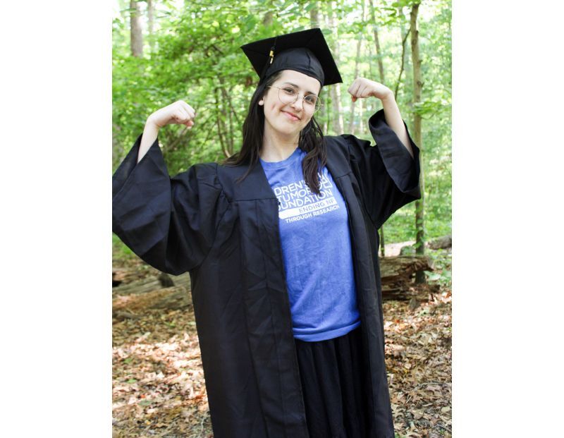 A person in a graduation cap and gown stands outdoors, smiling and flexing both arms, with trees and leaves in the background.